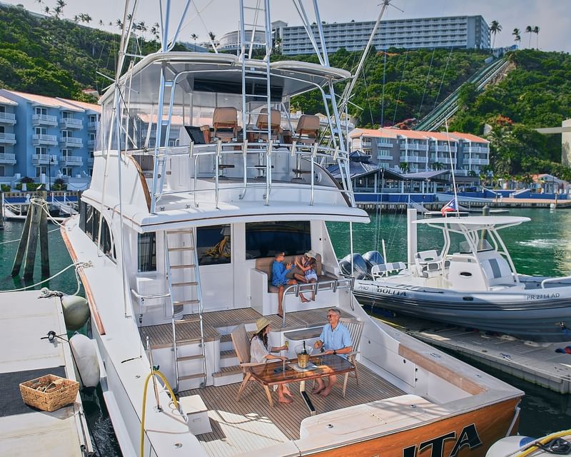 Family relaxing on the wooden deck of a luxury yacht docked at a marina in front of the hillside El Conquistador Resort