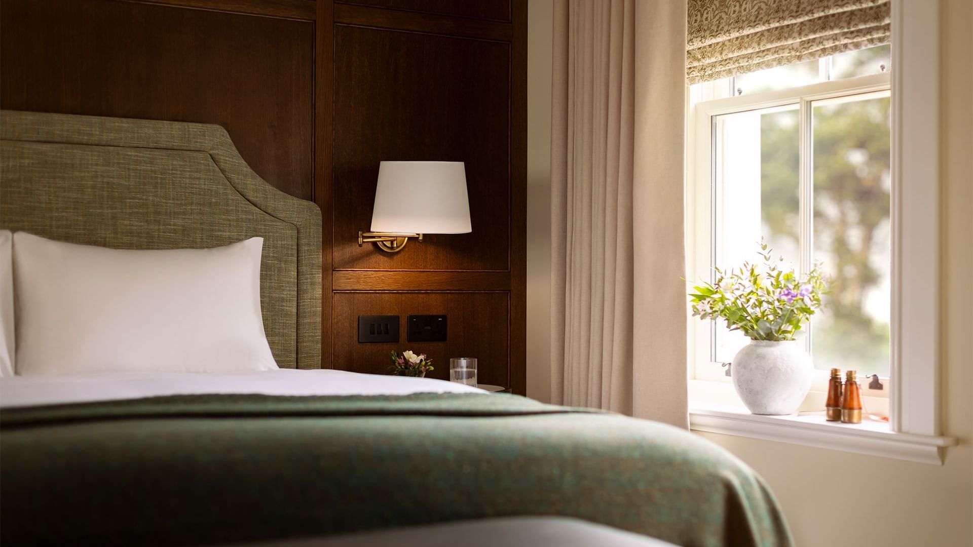 Bed with a lamp on the wall, and a vase of flowers on the windowsill in Twin Courtyard Suite at Dunluce Lodge