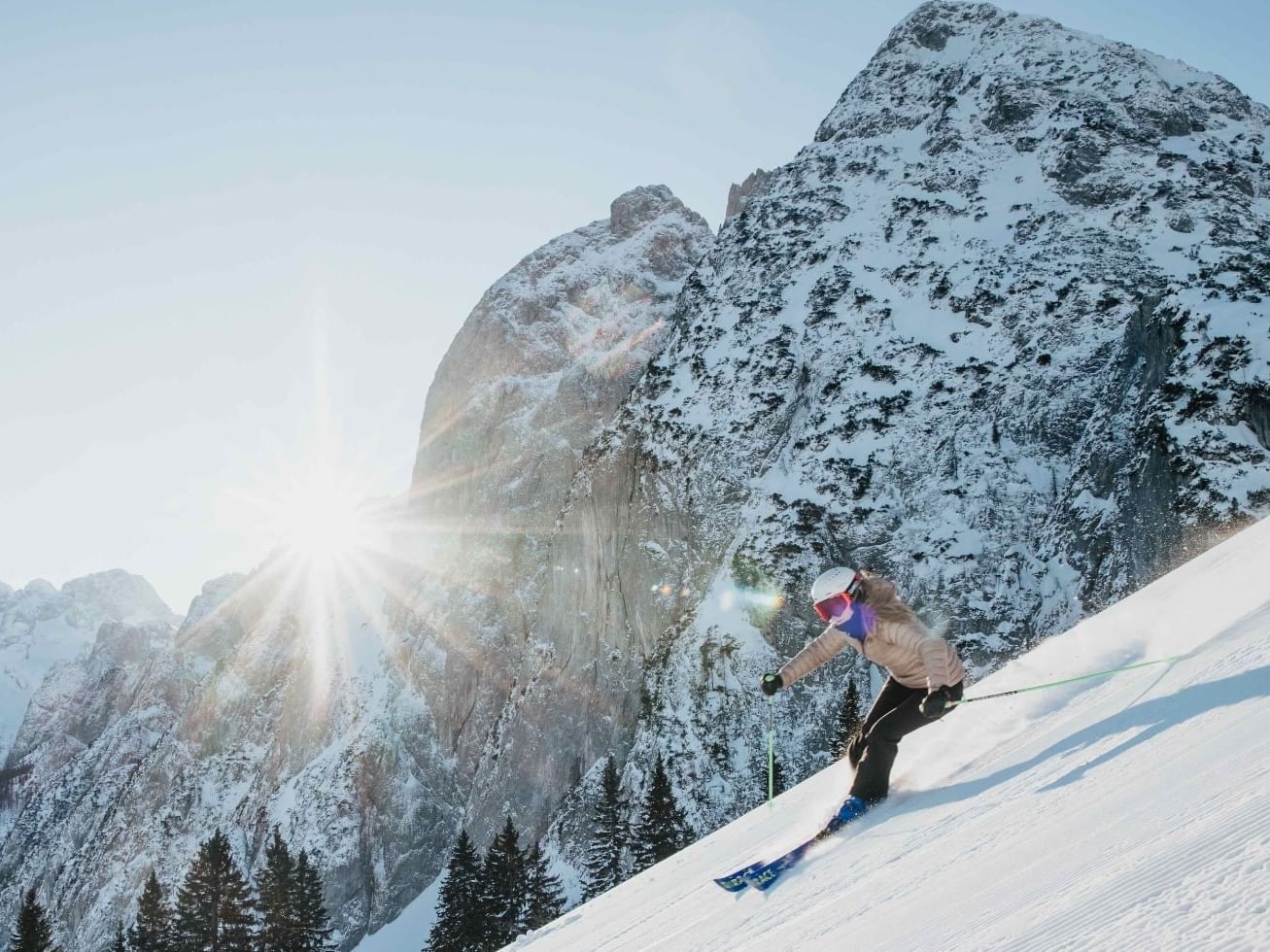 Skier in winter gear descends snowy mountain slope with sun shining brightly behind them.