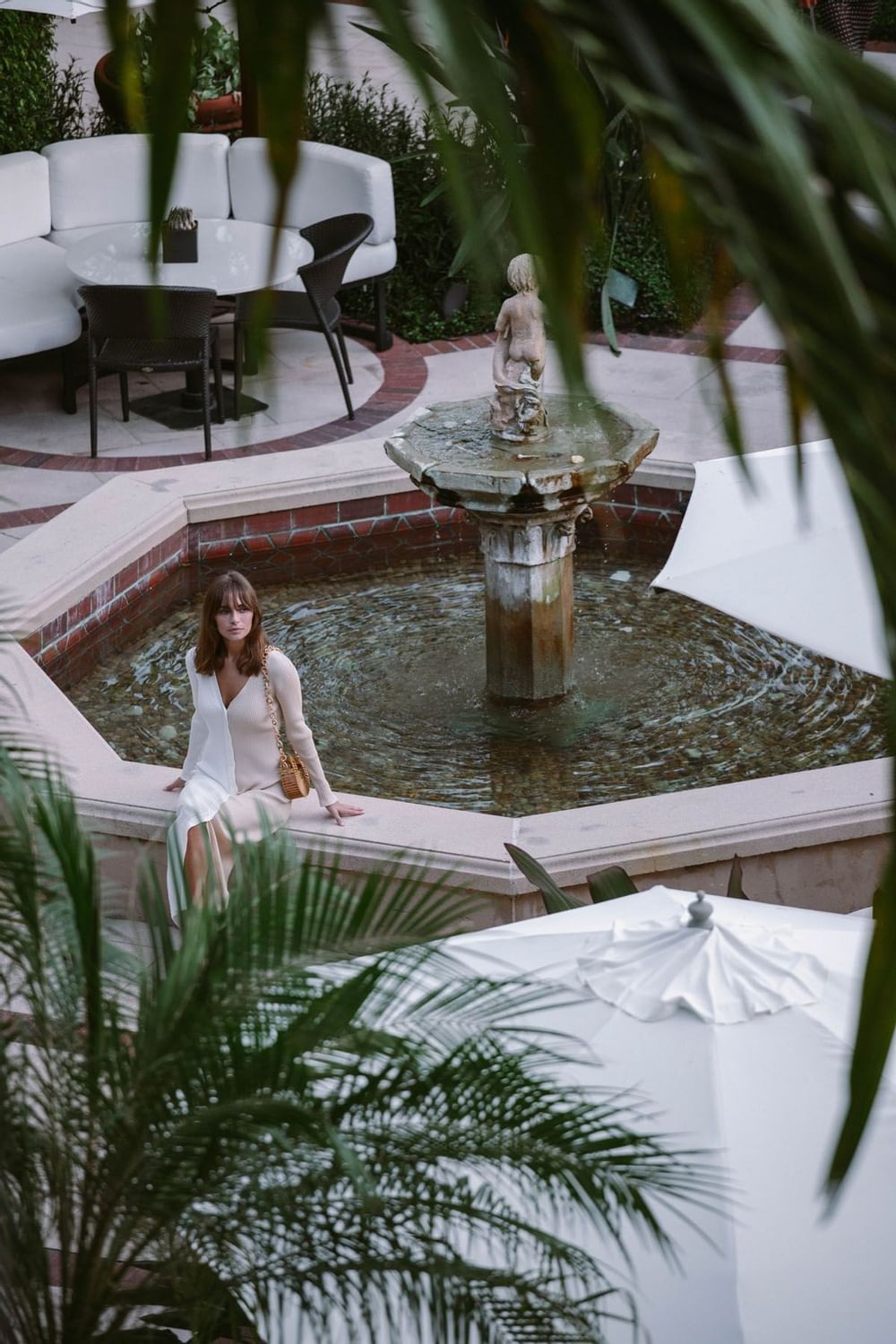 Woman in dress sitting on stone ledge of fountain surrounded by palm trees in courtyard pool at The Brazilian Court