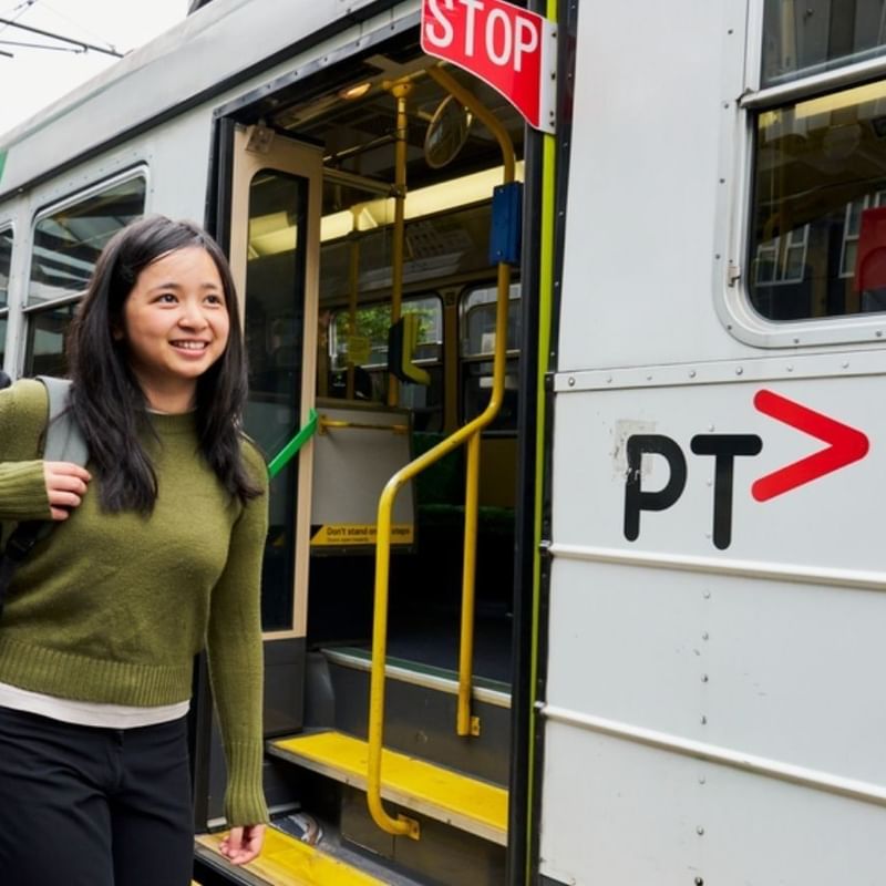 Smiling student with backpack boarding a public transit tram at an urban station platform for StudentLiving by UniLodge
