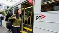 Smiling student with backpack boarding a public transit tram at an urban station platform for StudentLiving by UniLodge
