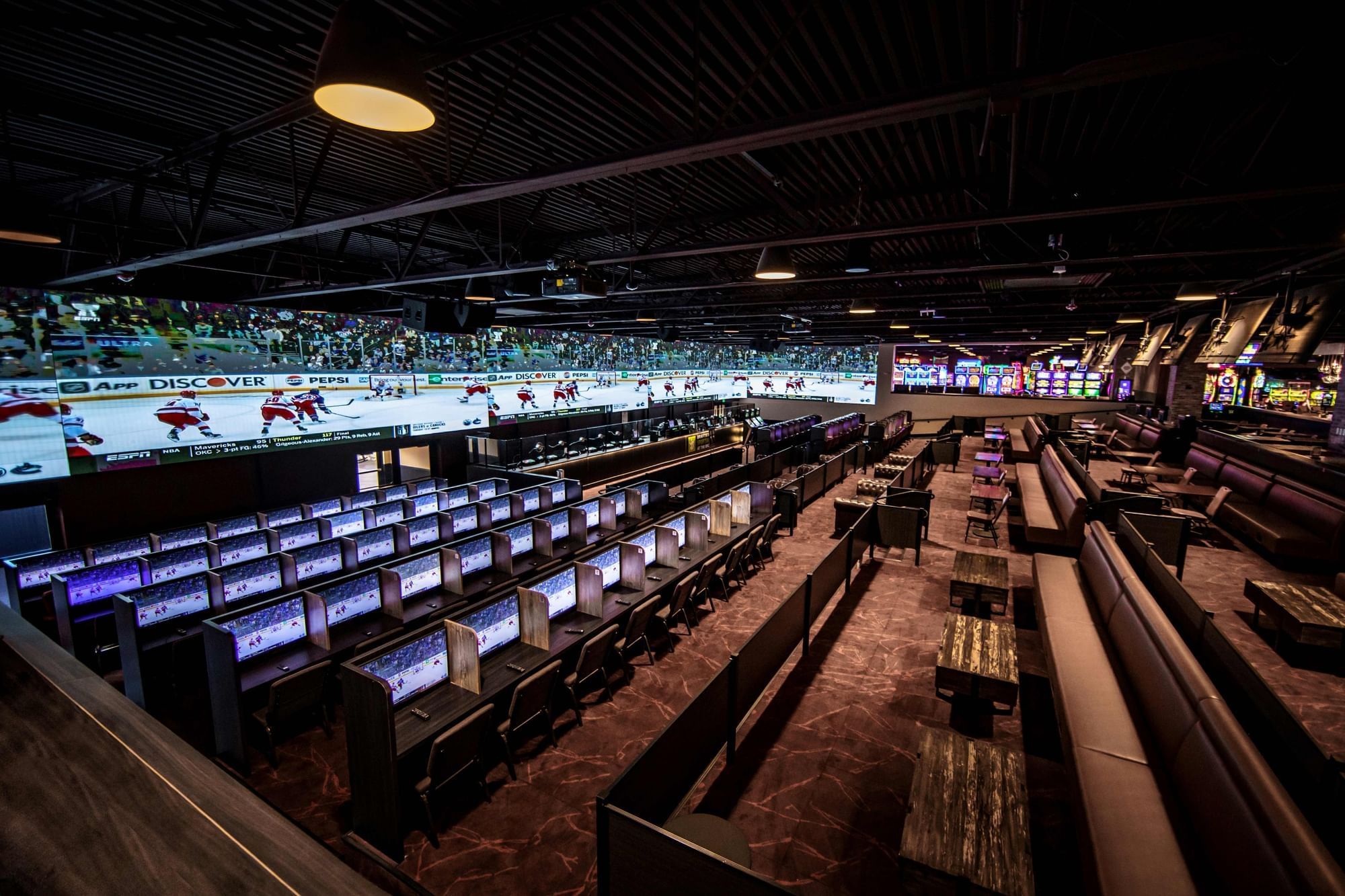 Sportsbook with betting screens and seats under hockey displays, beside lounge booths and tables at The Brook