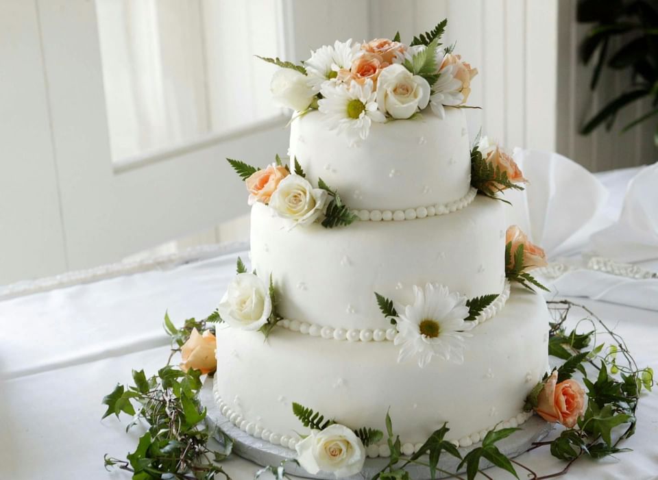 Three-tier white wedding cake with peach roses, daisies, and ferns, set on a linen-draped table at Ananea Madivaru Maldives