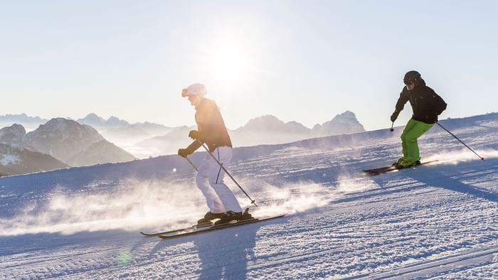 Due sciatori in discesa su una pista innevata, con montagne innevate sullo sfondo.