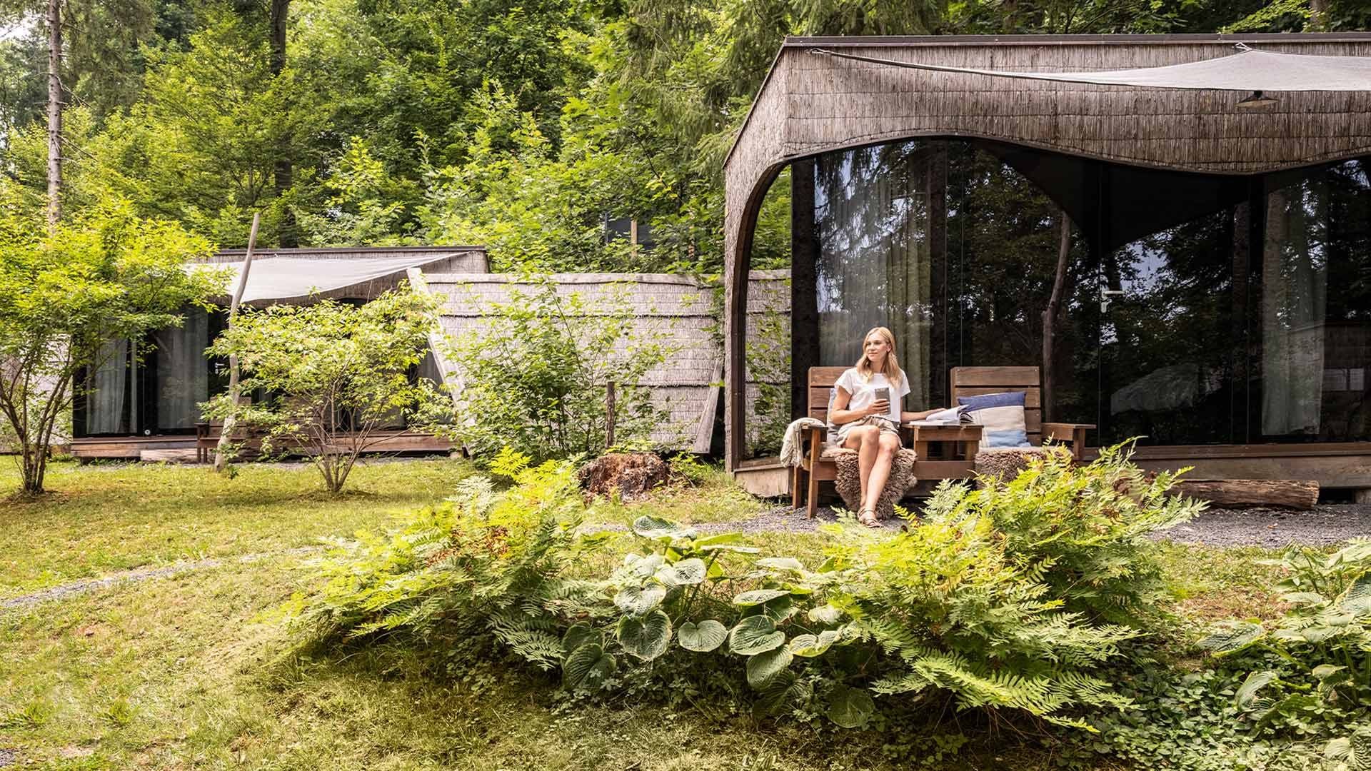 Woman sitting on a bench in front of a glass house with a lush garden and trees.