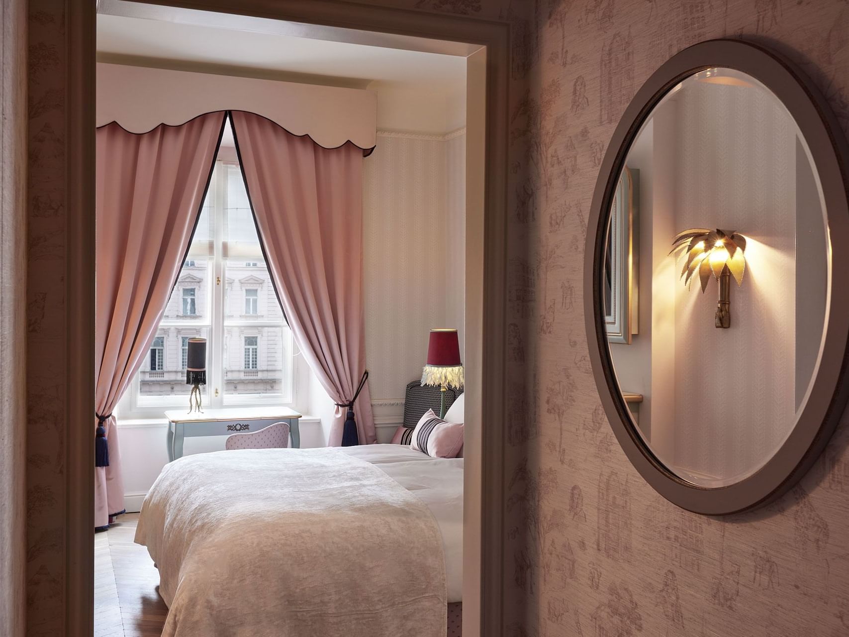 A bedroom with a bed, pink curtains, a red lamp, and a round mirror with a decorative light.