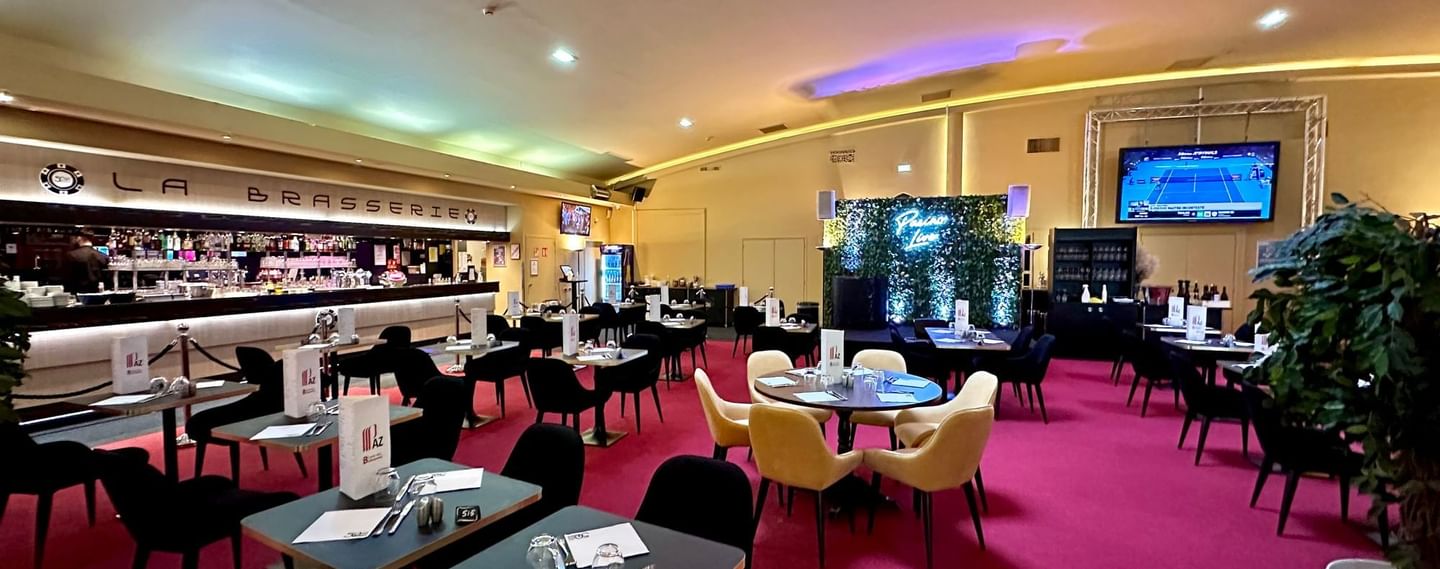 Salle de restaurant avec tables, chaises, moquette rouge, bar, écrans et comptoir de boissons.