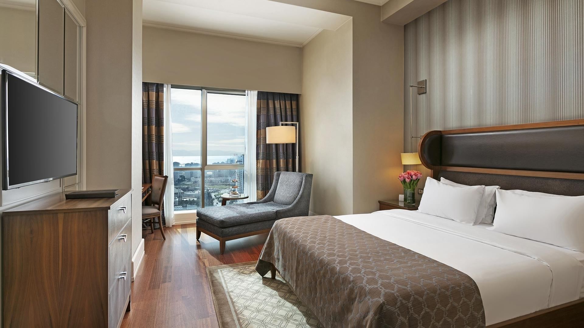 Titanic Business Kartal offers a luxurious room featuring a large bed, chaise lounge, TV, dresser, and a spacious window with stunning city views