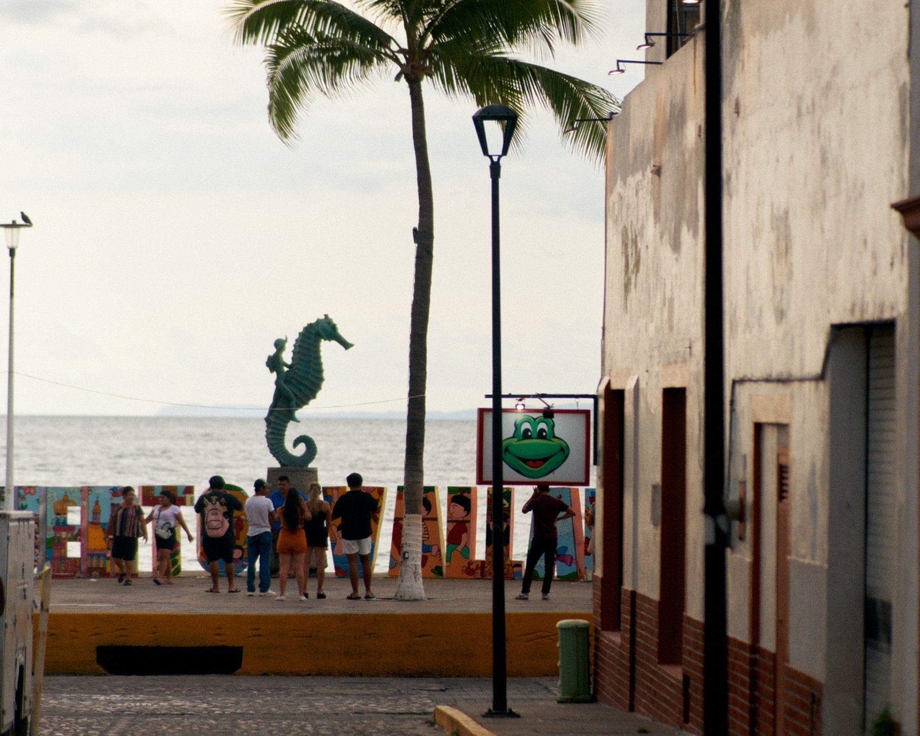 People walking along the Puerto Vallarta boardwalk near the Seahorse sculpture, palm trees, and a view of the Pacific Ocean.