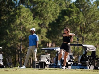 Woman in black athletic wear swinging golf club on sunny course with man and golf carts in background.