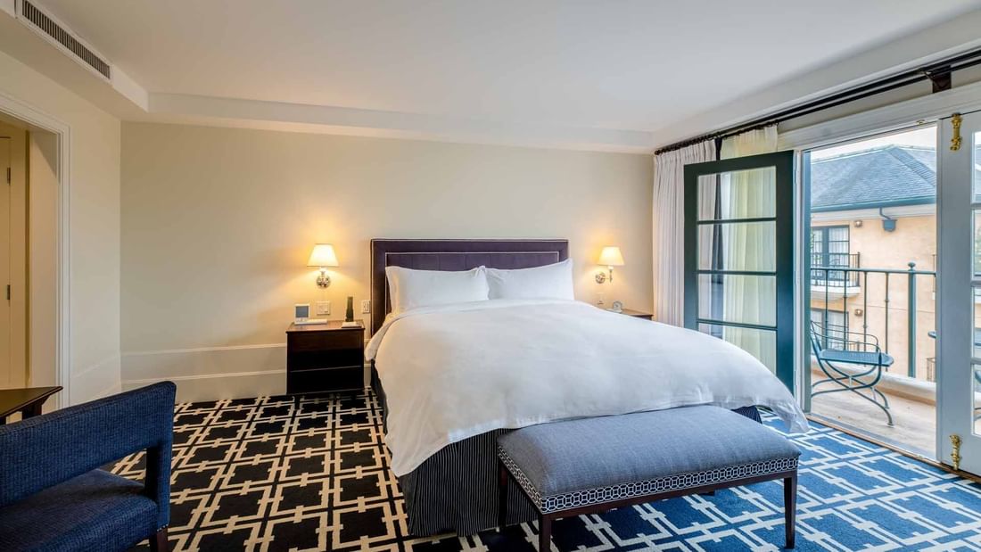 Seating area and ottoman by the cozy bed in Courtyard Deluxe Fireplace King with geometric patterned carpet at El Prado Hotel