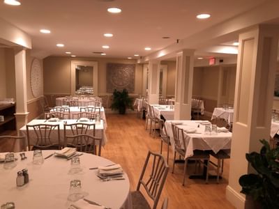 Round tables with white linens by wooden chairs, under recessed lighting near tapestry wall art at Simsbury 1820 House