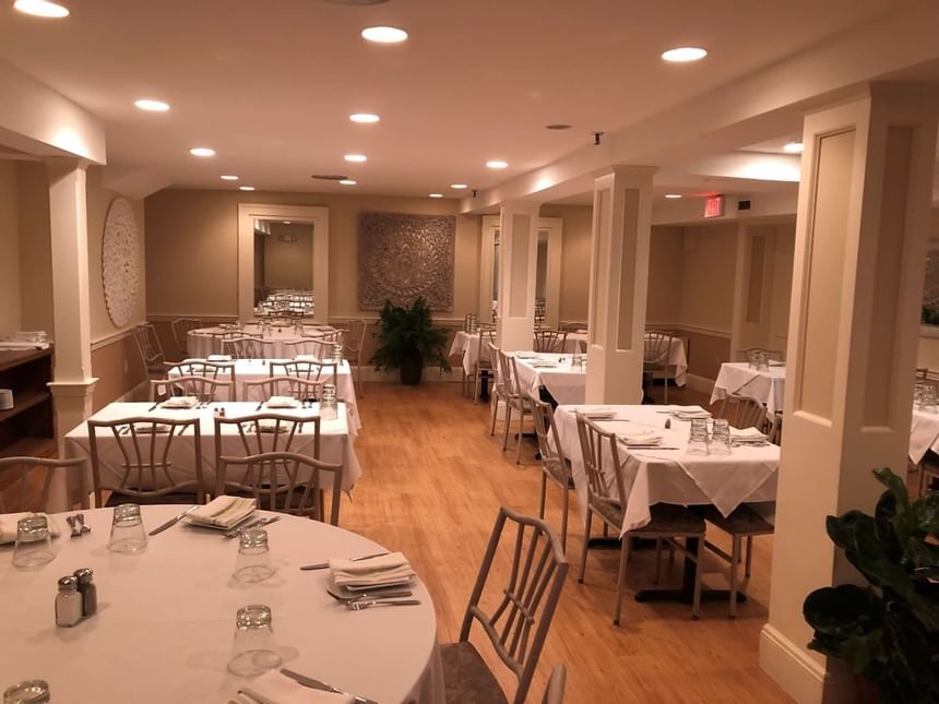 Round tables with white linens by wooden chairs, under recessed lighting near tapestry wall art at Simsbury 1820 House