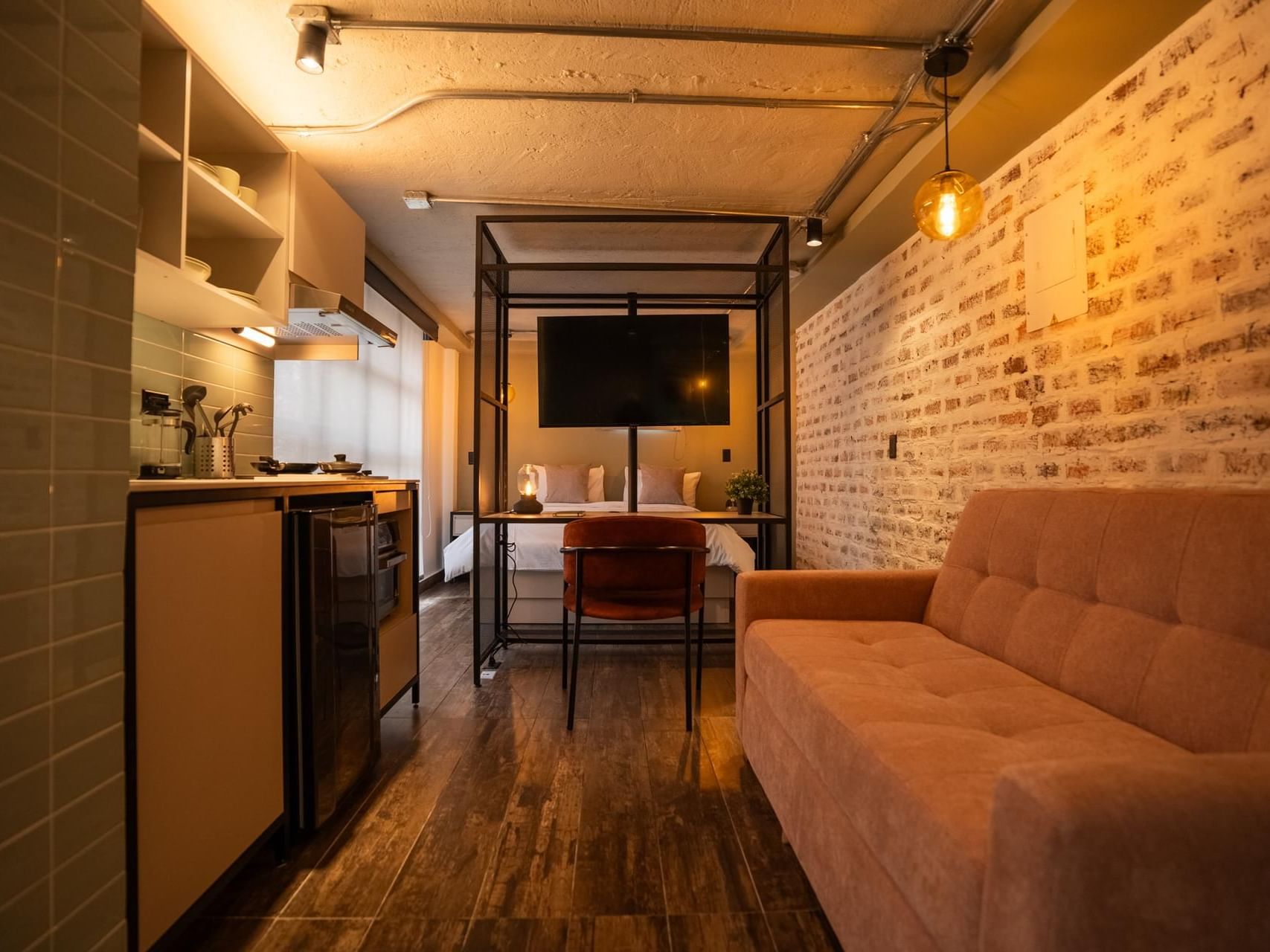 Estiloso loft con pared de ladrillo, sofá, cocina y cálidas luces doradas creando ambiente dentro de Studio Room en 5Q House Zona G
