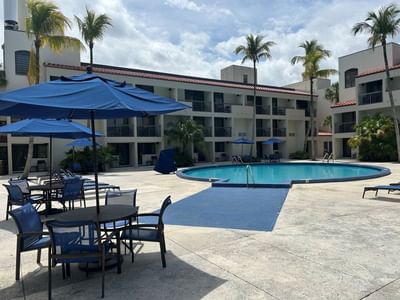 Outdoor swimming pool surrounded by palms and blue umbrellas near hotel balconies and sun loungers at Miami Lakes Hotel