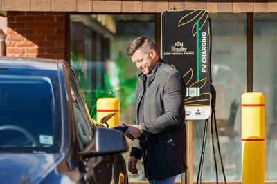 Smiling man in grey coat plugging a charging cable into an electric car at an EV station outside Rodd Royalty