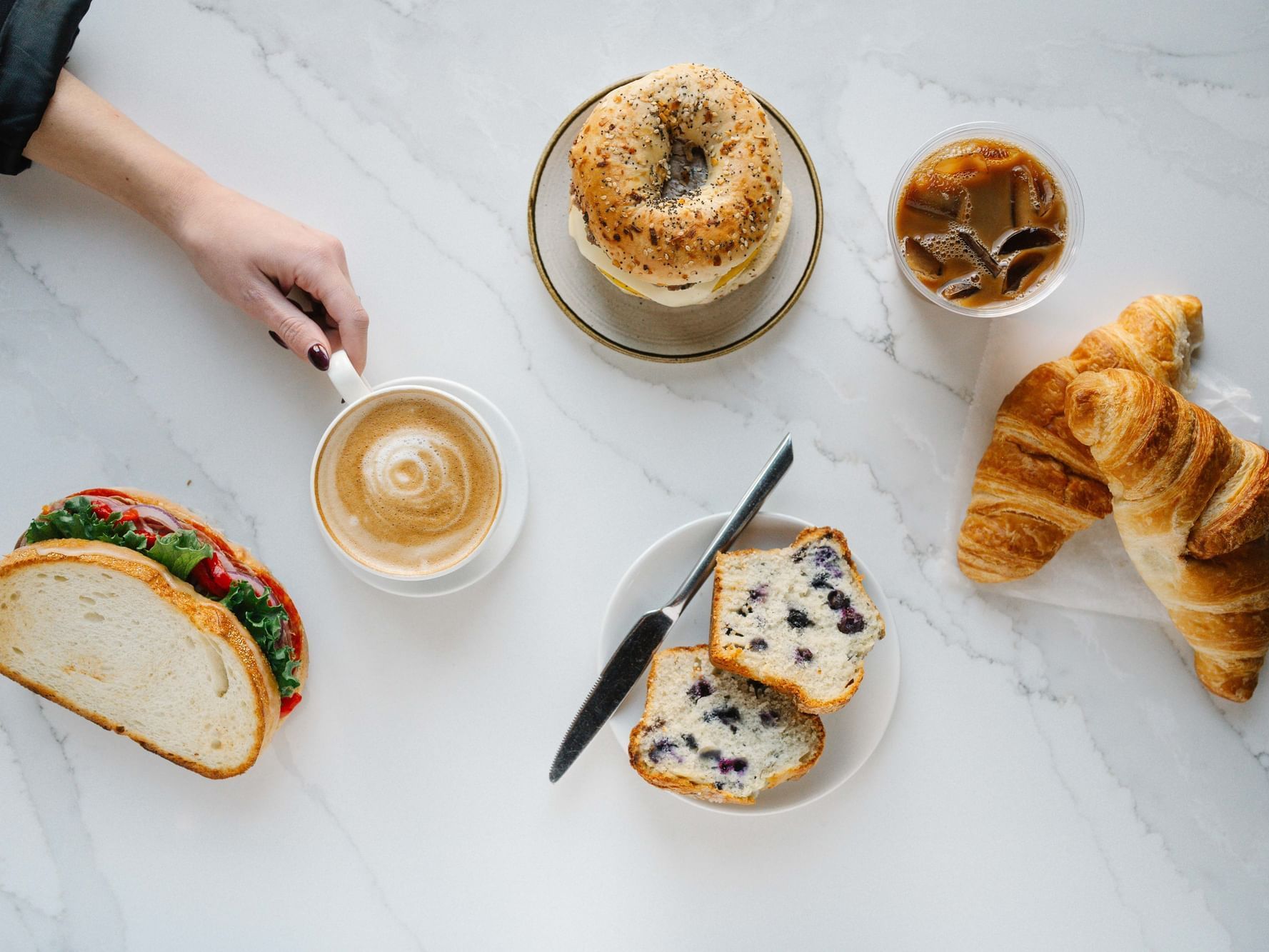 A breakfast spread with bagel, sandwich, croissant, blueberry bread, coffee, and iced coffee on a marble surface.