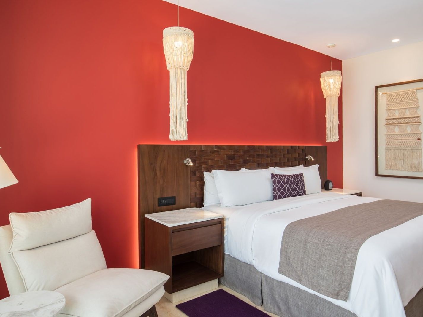 King Deluxe Bed with a vibrant red accent wall, macramé lamp, and a comfy white lounge chair at Camino Real Merida