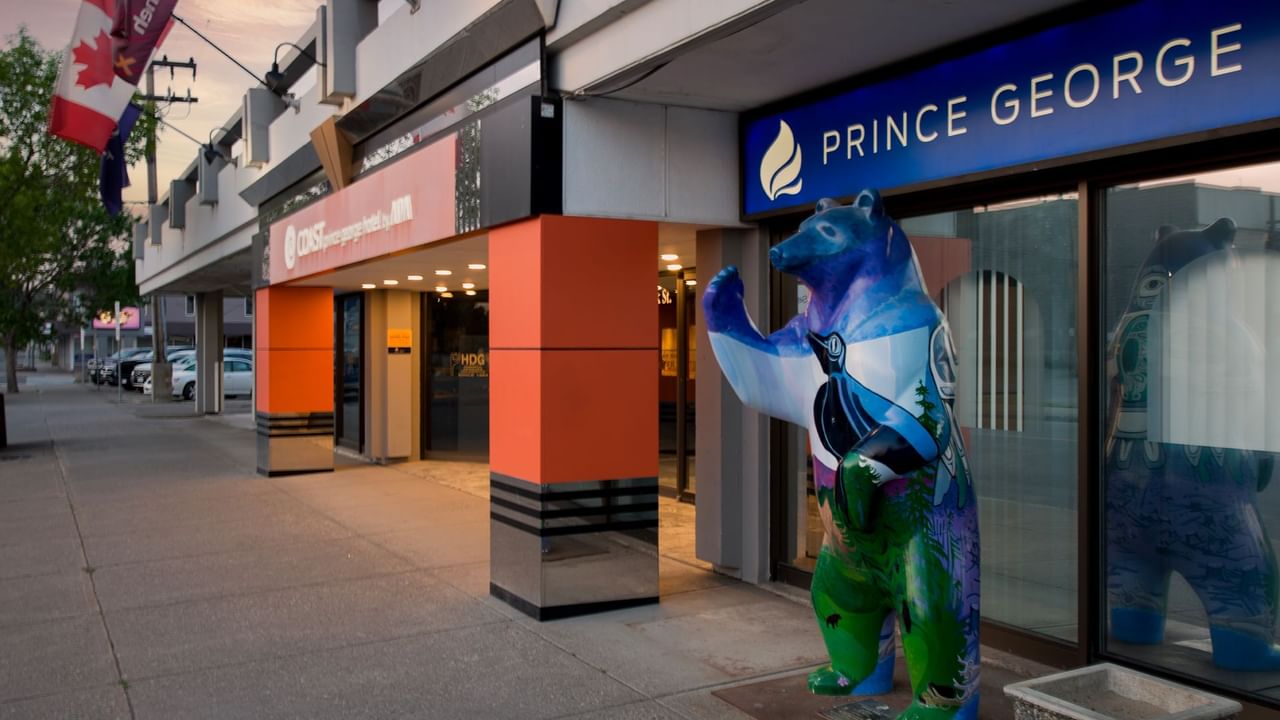 Entrance of Coast Prince George Hotel by APA with a colorful bear statue outside and a Canadian flag waving nearby.