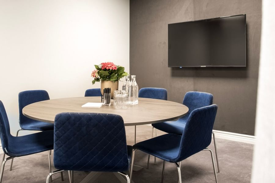 Round table with six blue chairs, a flower vase, and a flat-screen TV mounted on the wall.