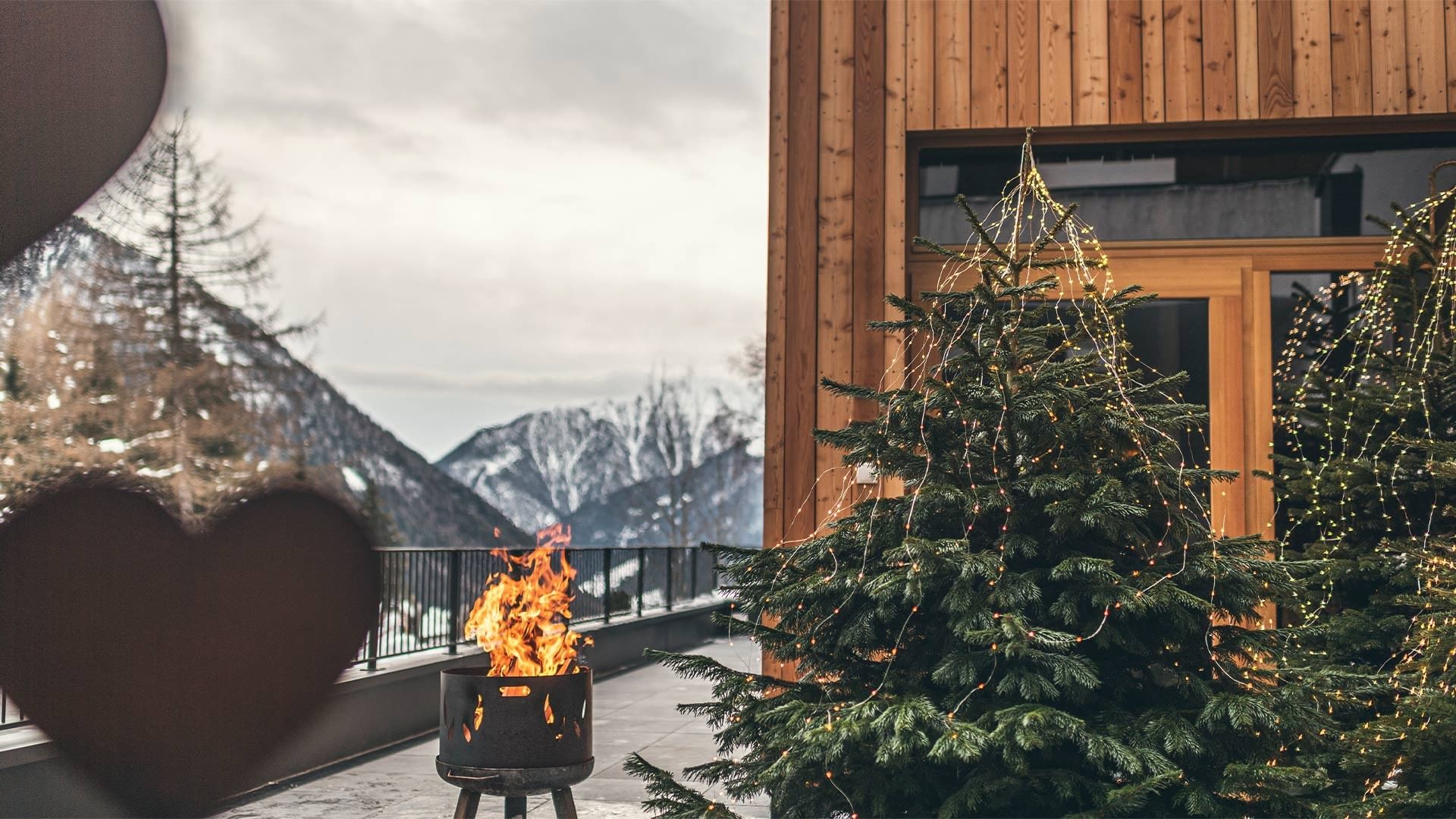 Feuerstelle und geschmückter Weihnachtsbaum vor einem Holzhaus mit Bergkulisse im Hintergrund.