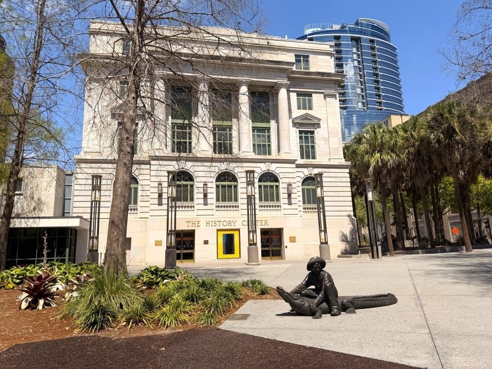 A large stone building with columns behind a garden walkway with a statue of a man wrangling an alligator. 