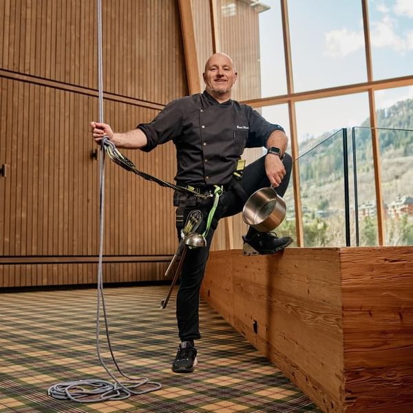 Ein Mann mit Kletterseilen und Kochutensilien, lächelnd neben einer Glaswand.