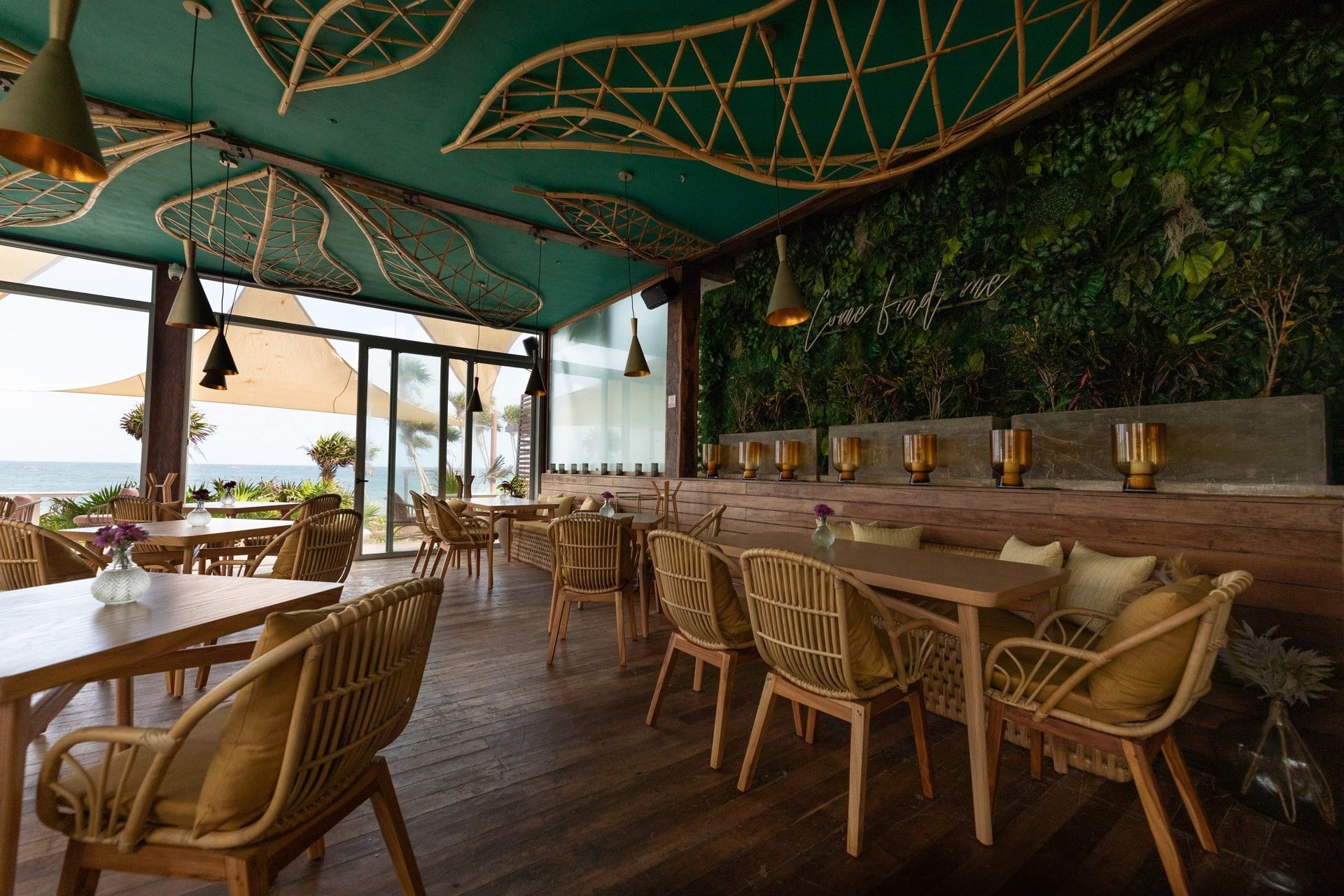 Mi Amor’s oceanfront dining room, with woven chairs, wooden tables and a green plant wall.