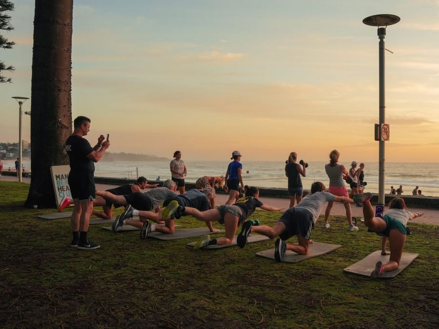 Yoga class at Elysium Manly, on mats under a sunset sky, surrounded by tall pine trees near a concrete walkway on the lawn