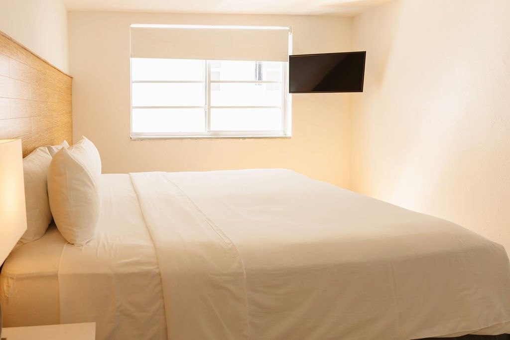 A neatly made king-sized bed with a wooden headboard, white linens, and pillows, facing a window with a wall-mounted TV to the right.