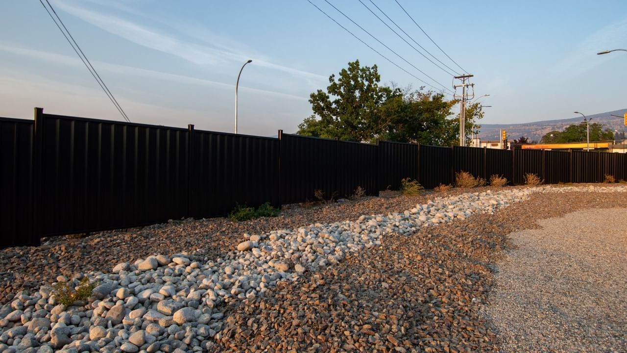 Black metal fence with gravel landscaping, trees, street lights, and power lines in the background.