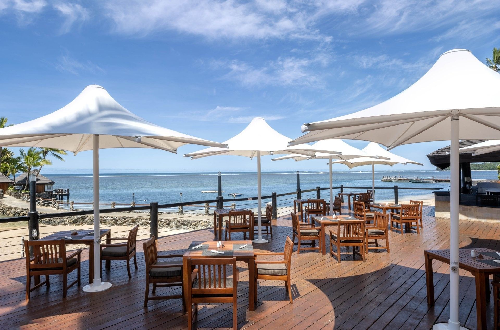 Wooden dining sets by a railing under white parasols surrounding a sunny deck at Warwick Fiji Resort and Spa