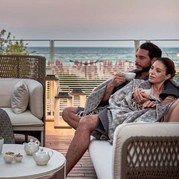 Couple wrapped in blankets, holding mugs, sitting on a balcony with ocean view.