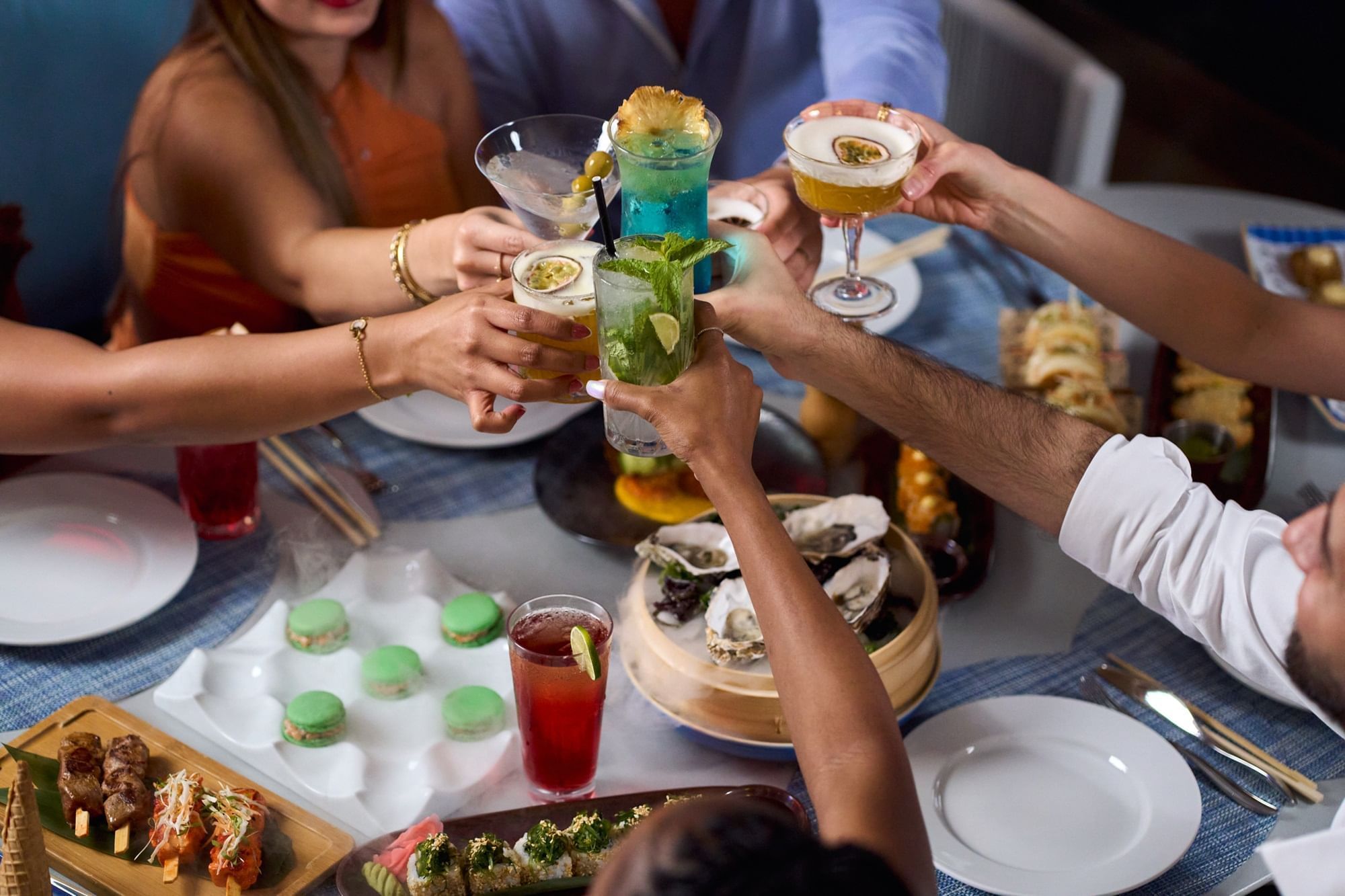 A group of friends cheers with colorful cocktails over a table of sushi, oysters, and desserts at Paramount Hotel Midtown.