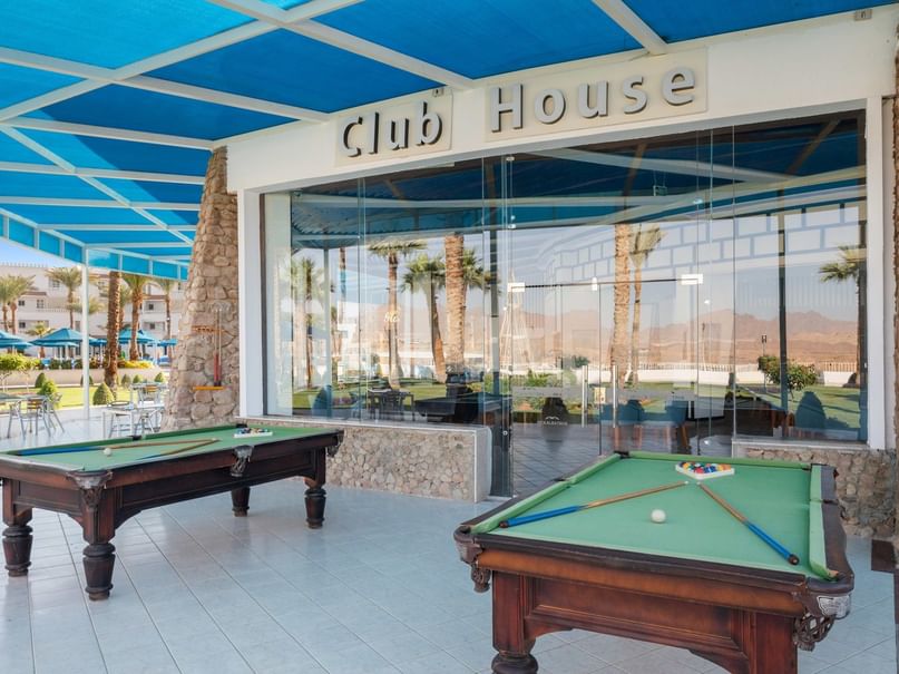 Two pool tables in front of a building named Club House with a blue canopy and glass walls.