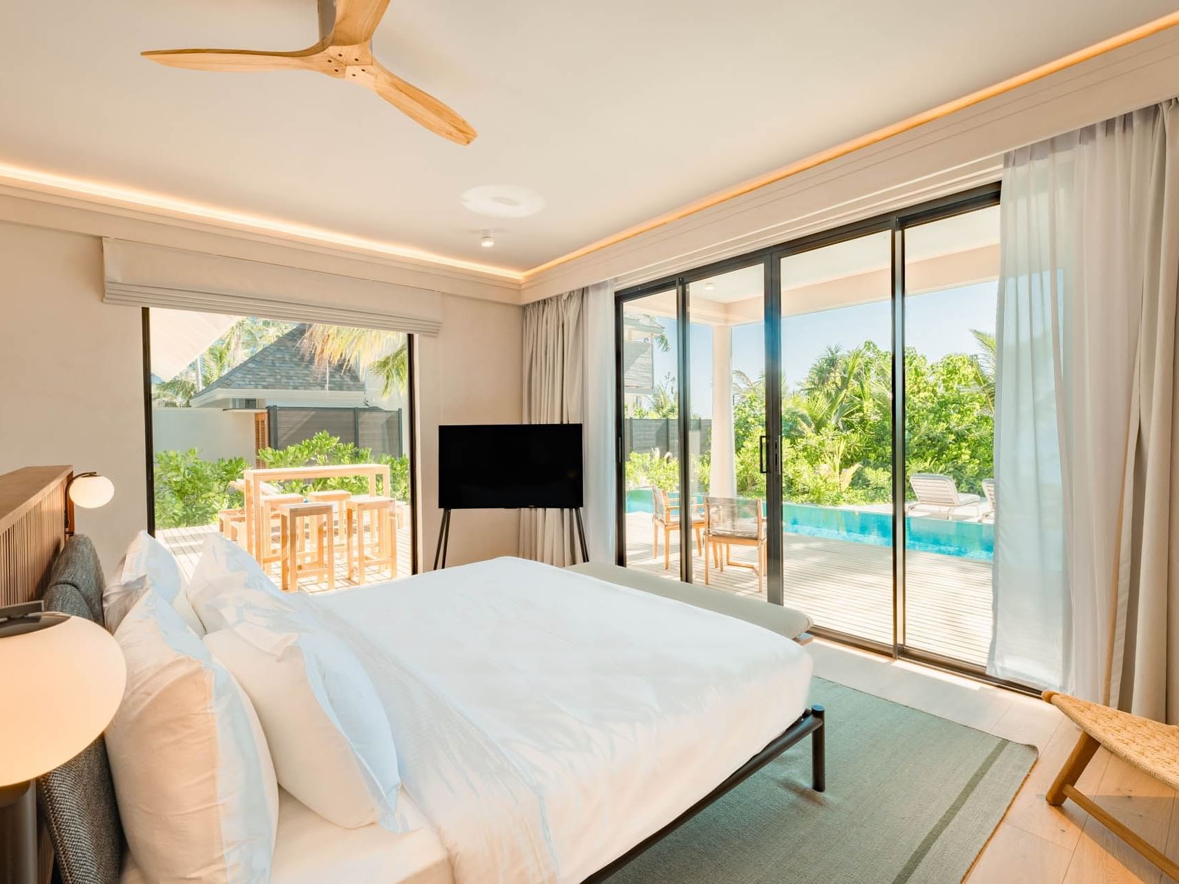 TV and ottoman by the bed in Beach Pool Villas with a view of lush trees and a pool outside at Ananea Madivaru Maldives