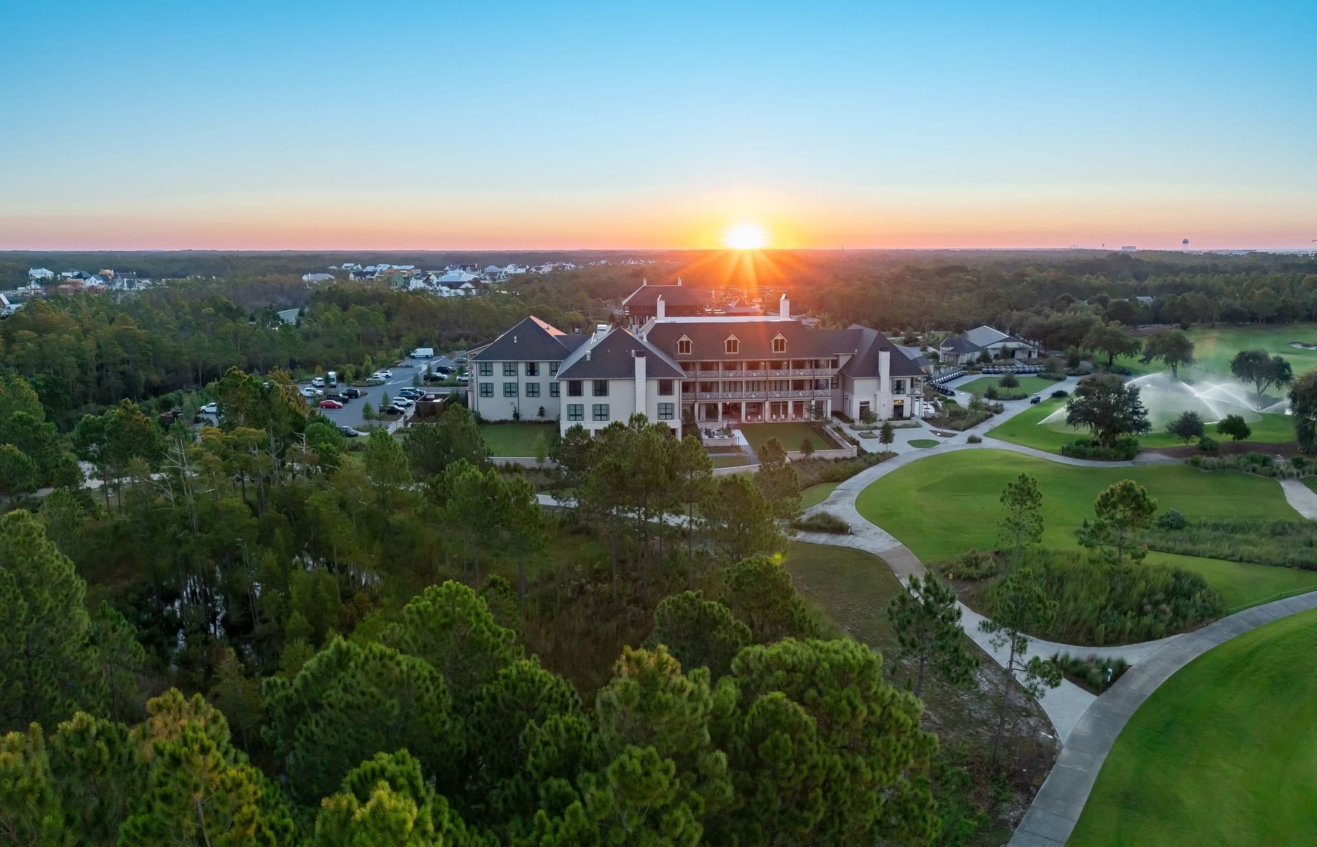 Aerial view of a large resort house surrounded by trees and golf courses, with the sun setting in the background.