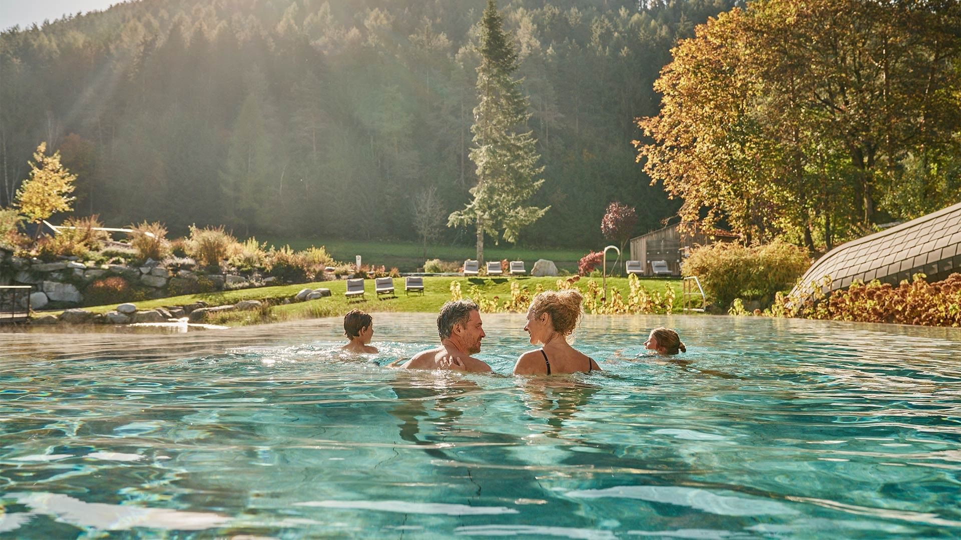 A family of four enjoys a pool with lush greenery, trees, and a distant mountain in the background.