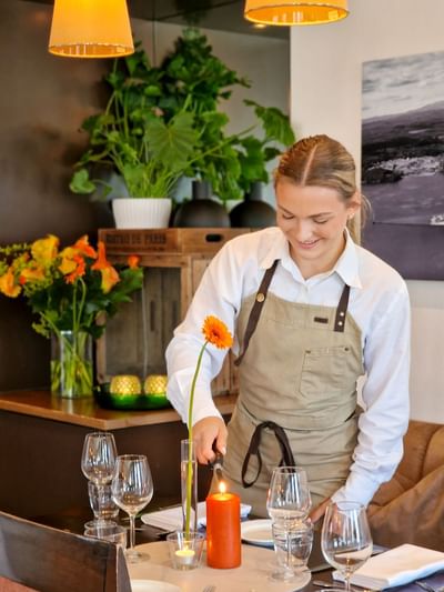 Smiling staff member lighting an orange candle on a dining table set with glassware and flowers at Tyrifjord Hotel in Norway