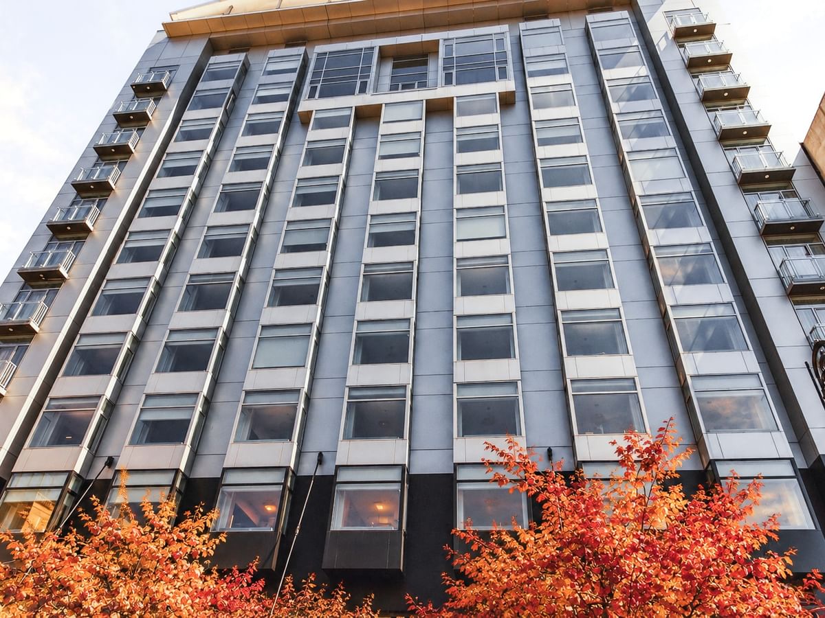 Modern hotel building with glass facade and balconies framed by vibrant orange autumn foliage in the foreground.