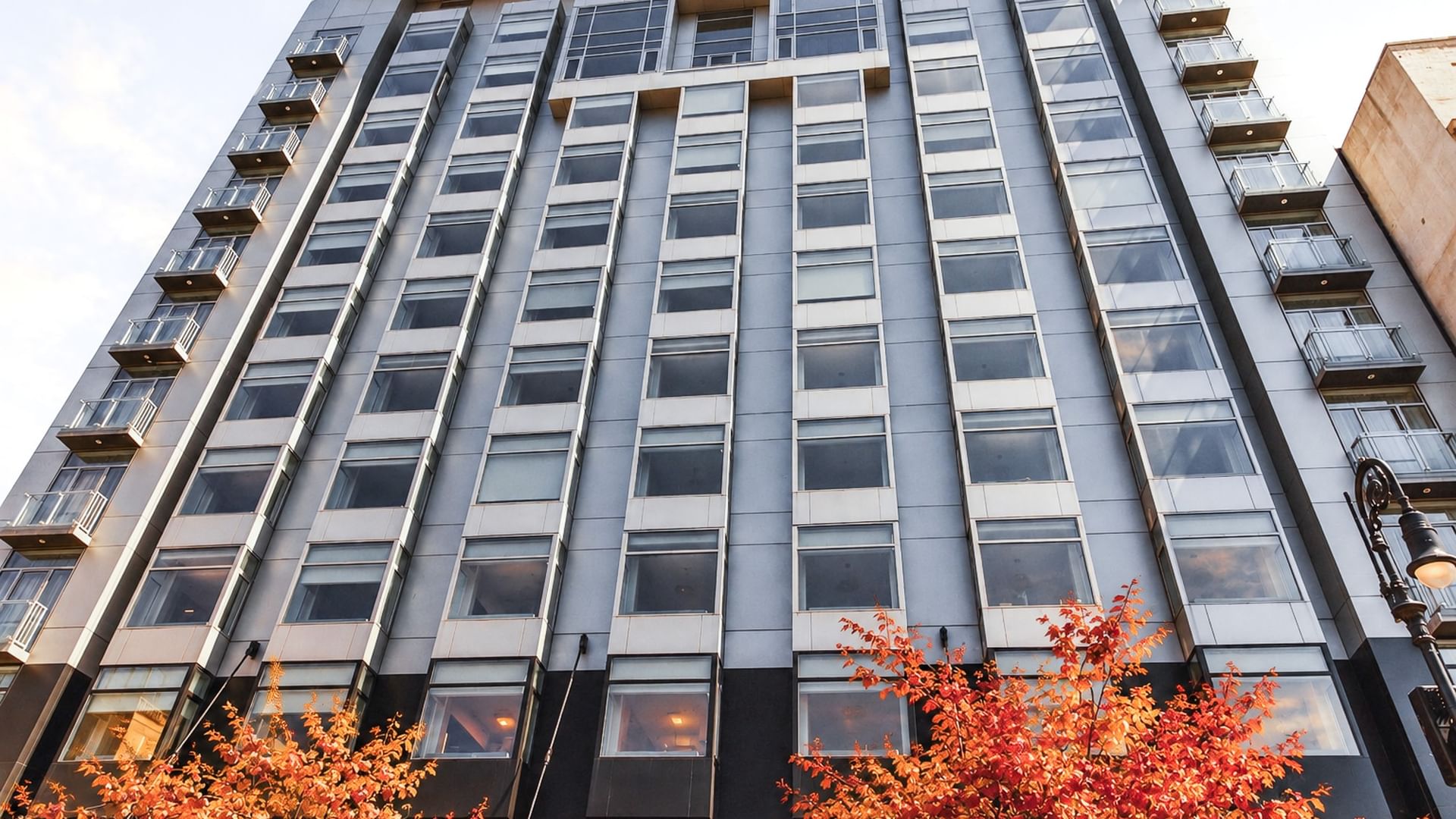 Modern hotel building with glass facade and balconies framed by vibrant orange autumn foliage in the foreground.
