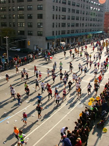 Crowd of runners moving along a city street by an urban park, surrounded by tall buildings near the Warwick New York