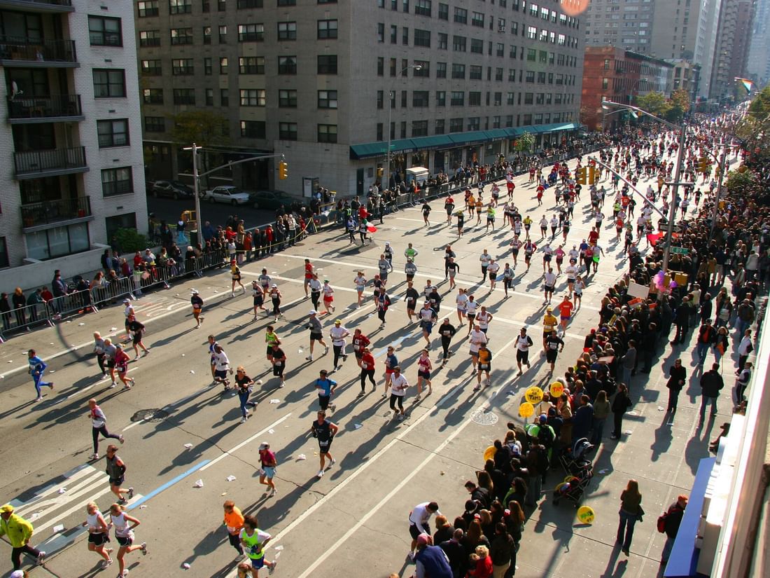 Crowd of runners moving along a city street by an urban park, surrounded by tall buildings near the Warwick New York
