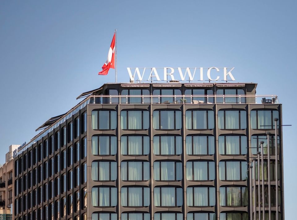Exterior View of Warwick Geneva hotel with flag and rooftop signage under clear sky