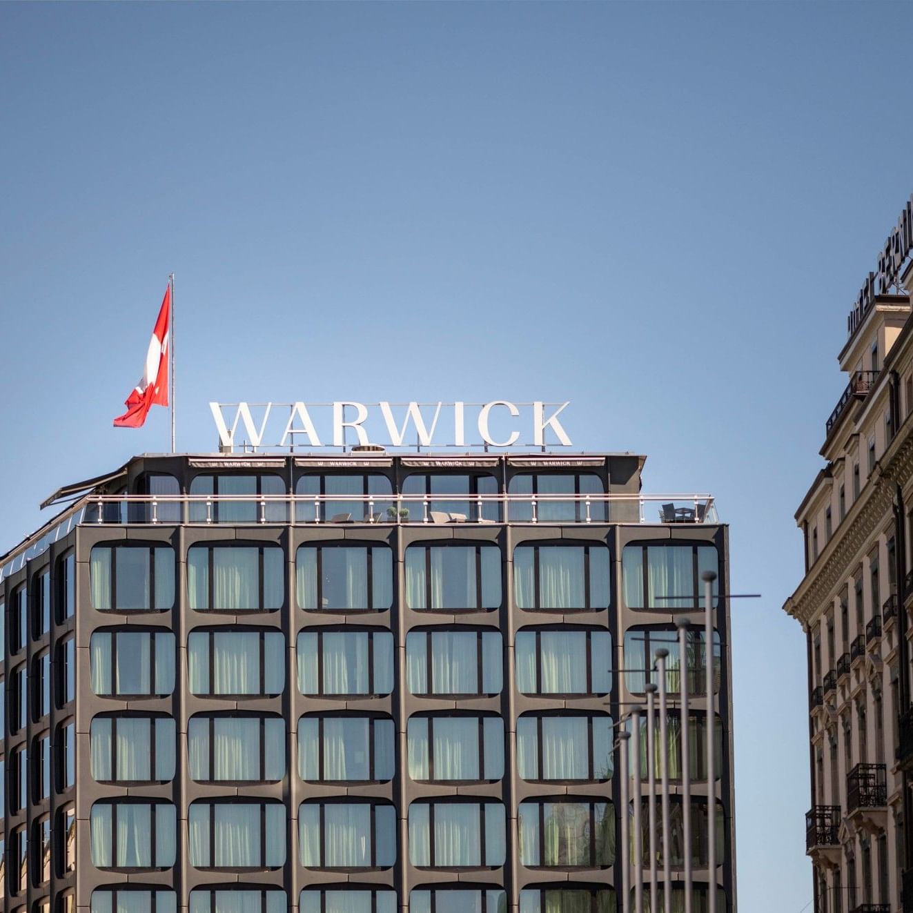 Extérieur de l'hôtel Warwick Geneva, avec son drapeau et son enseigne sur le toit, par une journée ensoleillée.