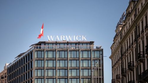 Extérieur de l'hôtel Warwick Geneva, avec son drapeau et son enseigne sur le toit, par une journée ensoleillée.