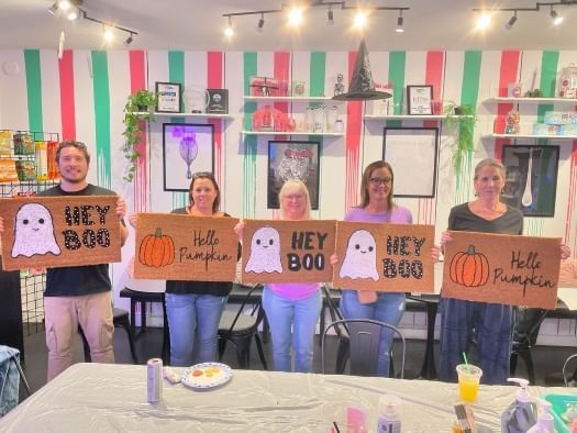 5 people stand in an art studio displaying handmade Halloween-themed doormats with pumpkins and ghosts. 