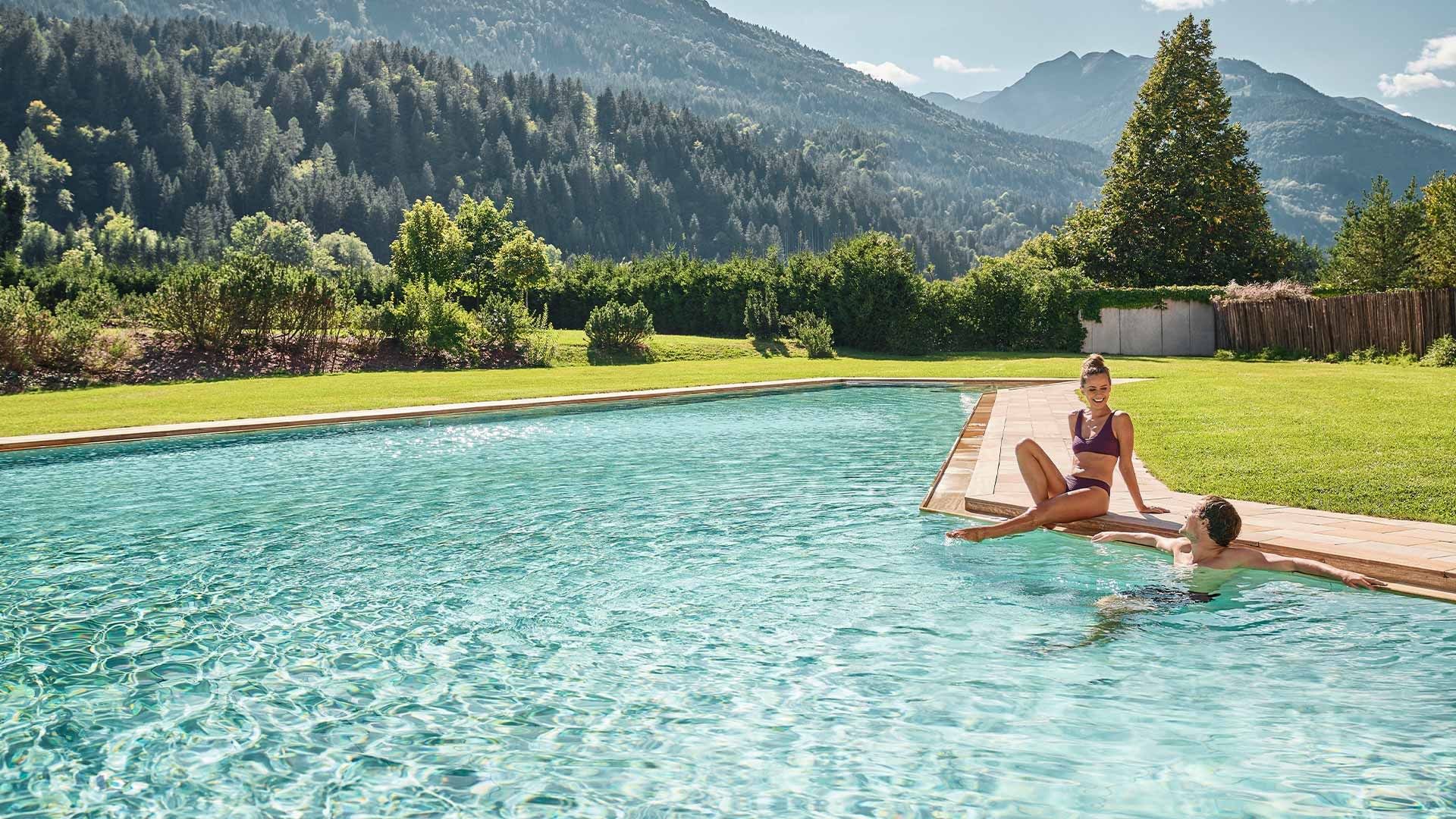 Donna e uomo godono di una piscina all'aperto circondata da prati e montagne.
