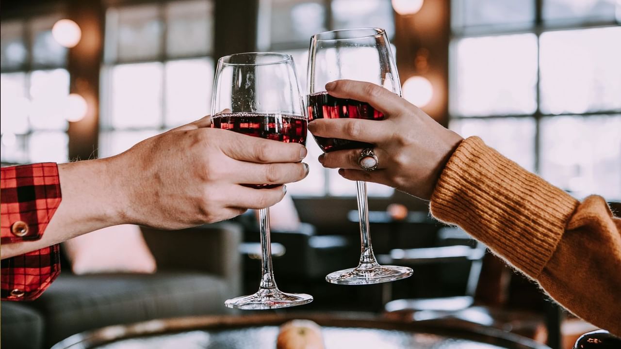 Two people clinking glasses of red wine, seated at a table in a restaurant setting.